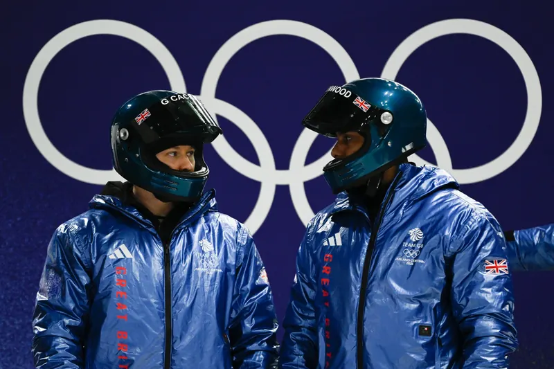 Leon Greenwood and a Team GB bobsleigh teammate in helmets and team jackets beneath the Olympic rings